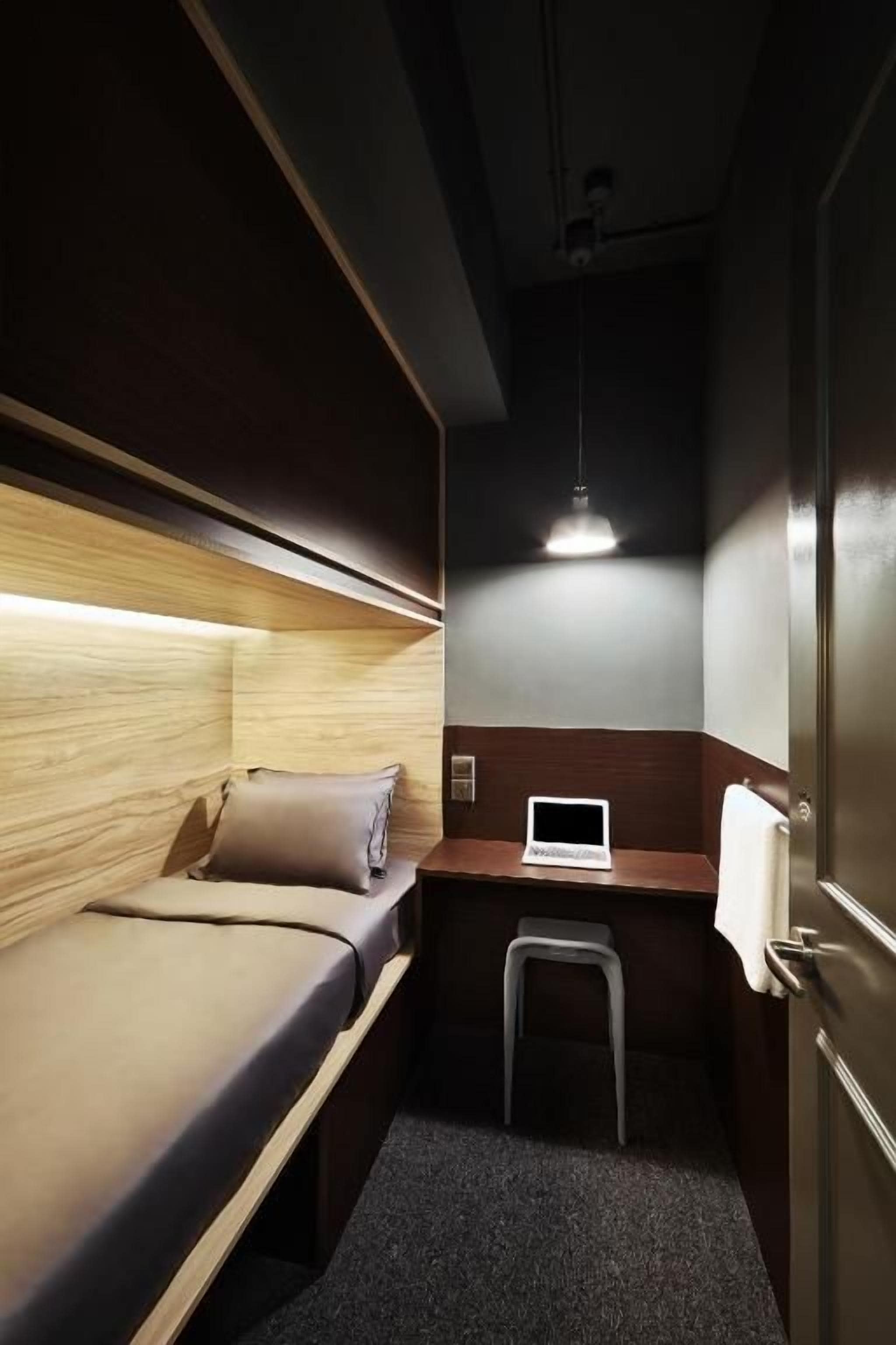 Ein kleines Zimmer mit einem Bett, einem Schreibtisch, einem Laptop und einer Pendelleuchte.