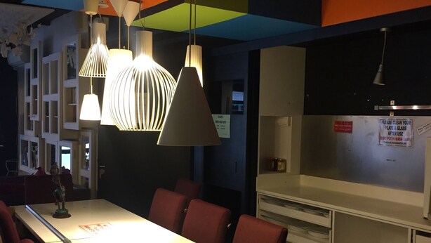 Un espace repas moderne avec une longue table, des chaises rouges et des luminaires suspendus. Des panneaux indiquent qu’un petit-déjeuner est servi quotidiennement et demandent aux clients de nettoyer la table et les verres après utilisation.