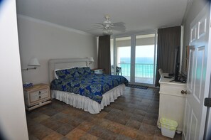 A hotel room with a bed, a nightstand, a desk, a ceiling fan, a balcony with a view of the ocean, and a trash bin. - Direct Beachfront condo;Spacious balcony;Stunning unobstructed Oceanfront Views (Navarre)