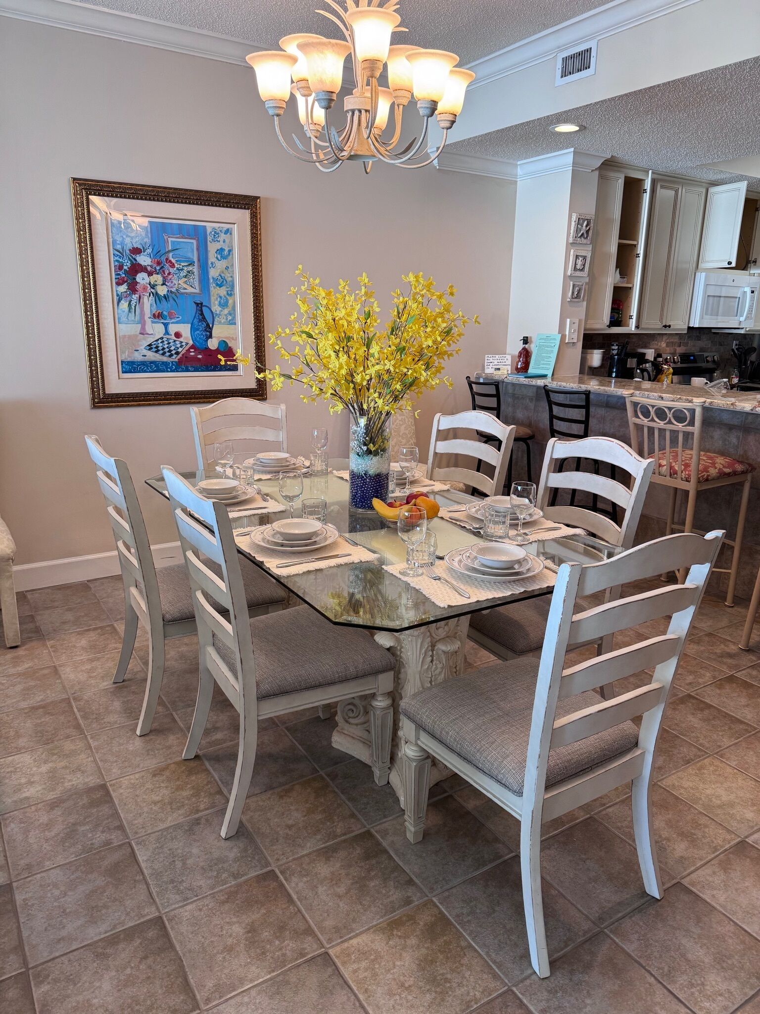 Comedor con una mesa de vidrio dispuesta para seis personas, rodeada por seis sillas. Un jarrón con flores amarillas está sobre la mesa.