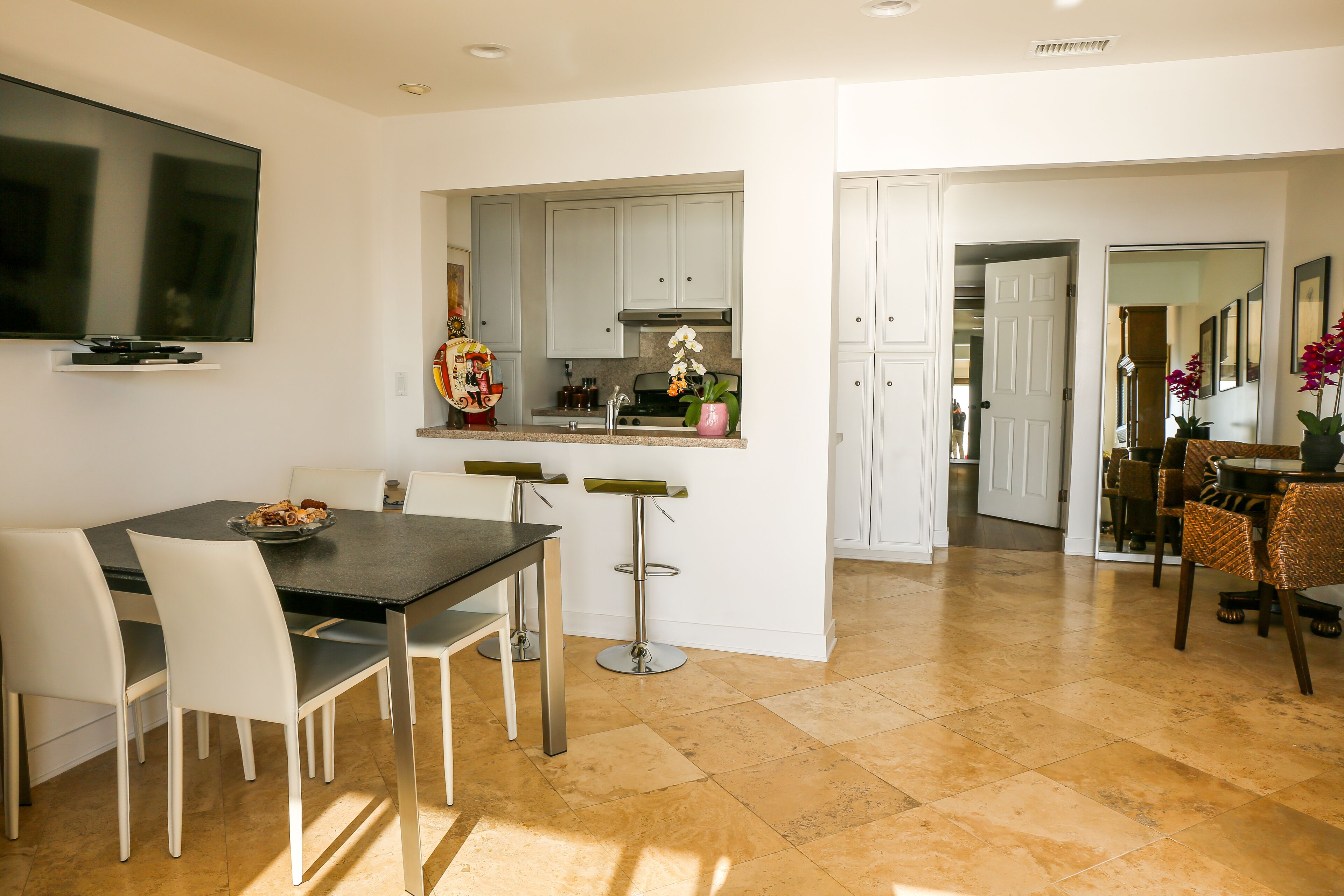 Una cocina moderna con armarios blancos, un comedor con una mesa oscura y sillas blancas, y un televisor montado en la pared.