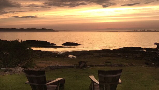 Une vue sur le coucher de soleil au-dessus d’une étendue d’eau, avec deux chaises en bois tournées vers l’horizon.