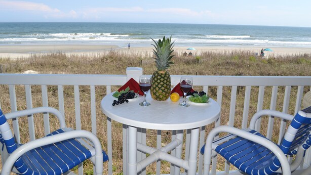 Une terrasse avec deux chaises à rayures bleues et une petite table sur laquelle sont disposés un ananas, des raisins et des verres à vin.