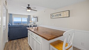 Une cuisine compacte avec un îlot central en bois, des meubles blancs et un ventilateur de plafond. Un panneau encadré accroché au mur porte l’inscription « SEASIDE ».