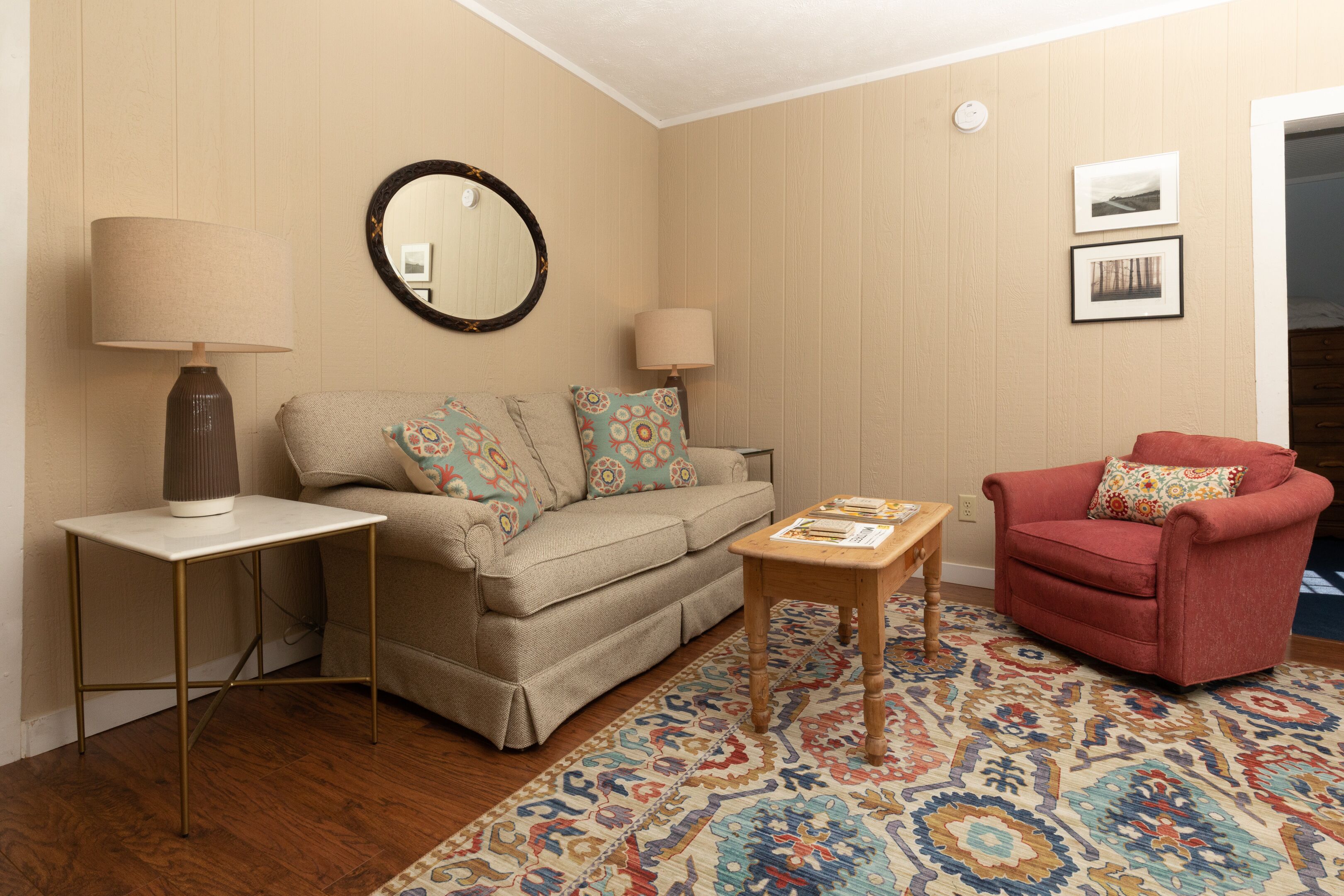 Una sala de estar con un sofá color crema, un sillón rojo, una mesa de centro, una mesita auxiliar, una lámpara, un espejo y un tapete estampado.
