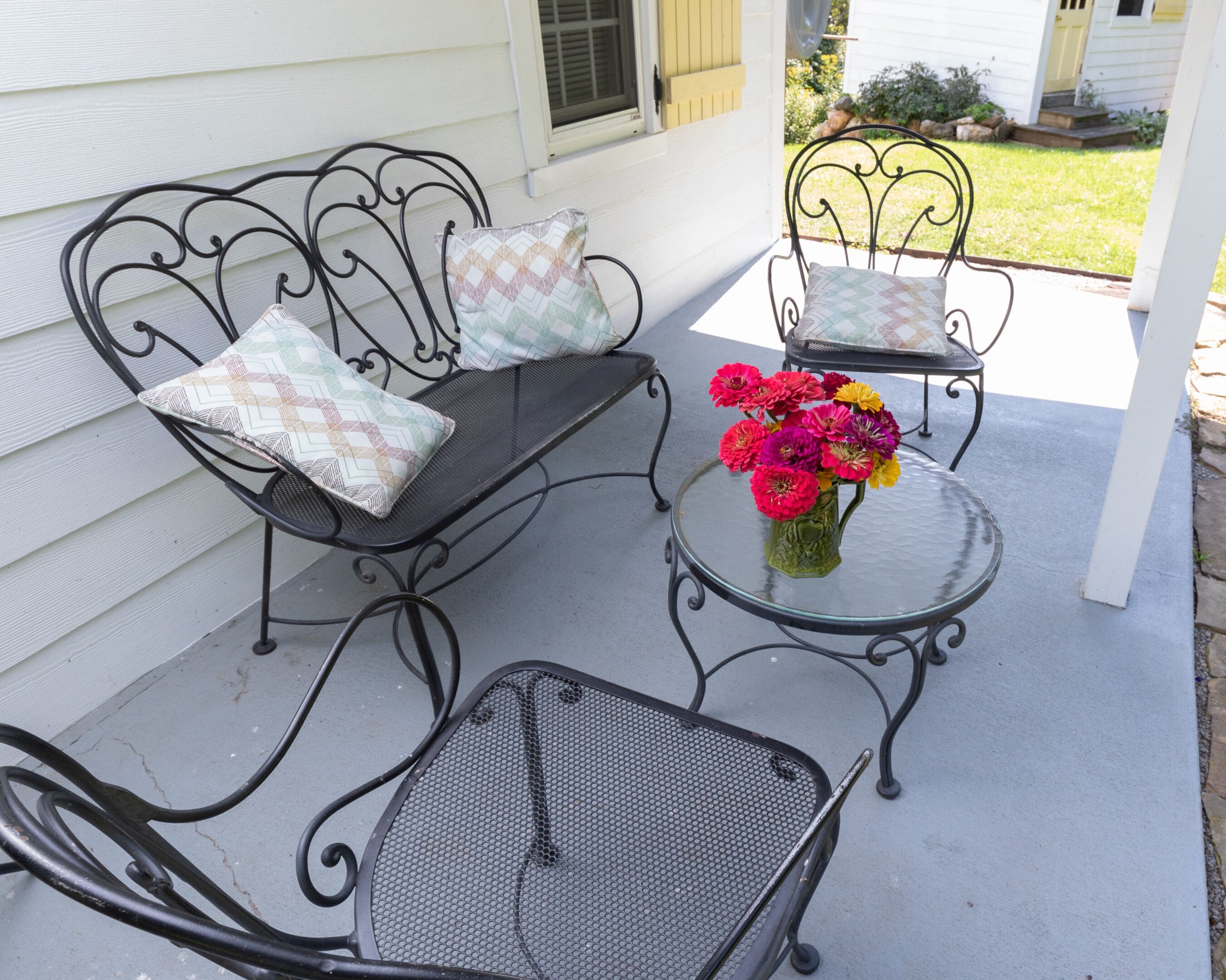 Een terras met twee metalen stoelen en een bank, elk voorzien van gekleurde, gemêleerde kussens. Een klein tafeltje met een vaas vol kleurrijke bloemen.