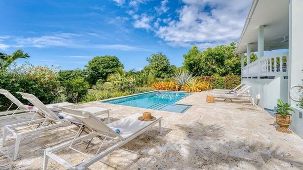 Un área de piscina con sillones de descanso, un cielo azul despejado y exuberante vegetación.