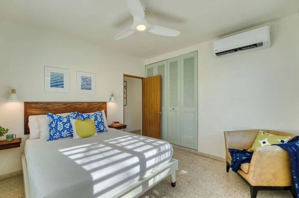 Une chambre d’hôtel comprenant un lit, un ventilateur de plafond, un appareil de climatisation, une porte en bois et un petit coin salon avec une chaise et des coussins.