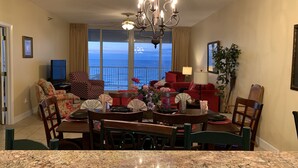 A spacious living room with a large dining table, comfortable seating, a chandelier, and a view of the ocean through large windows.