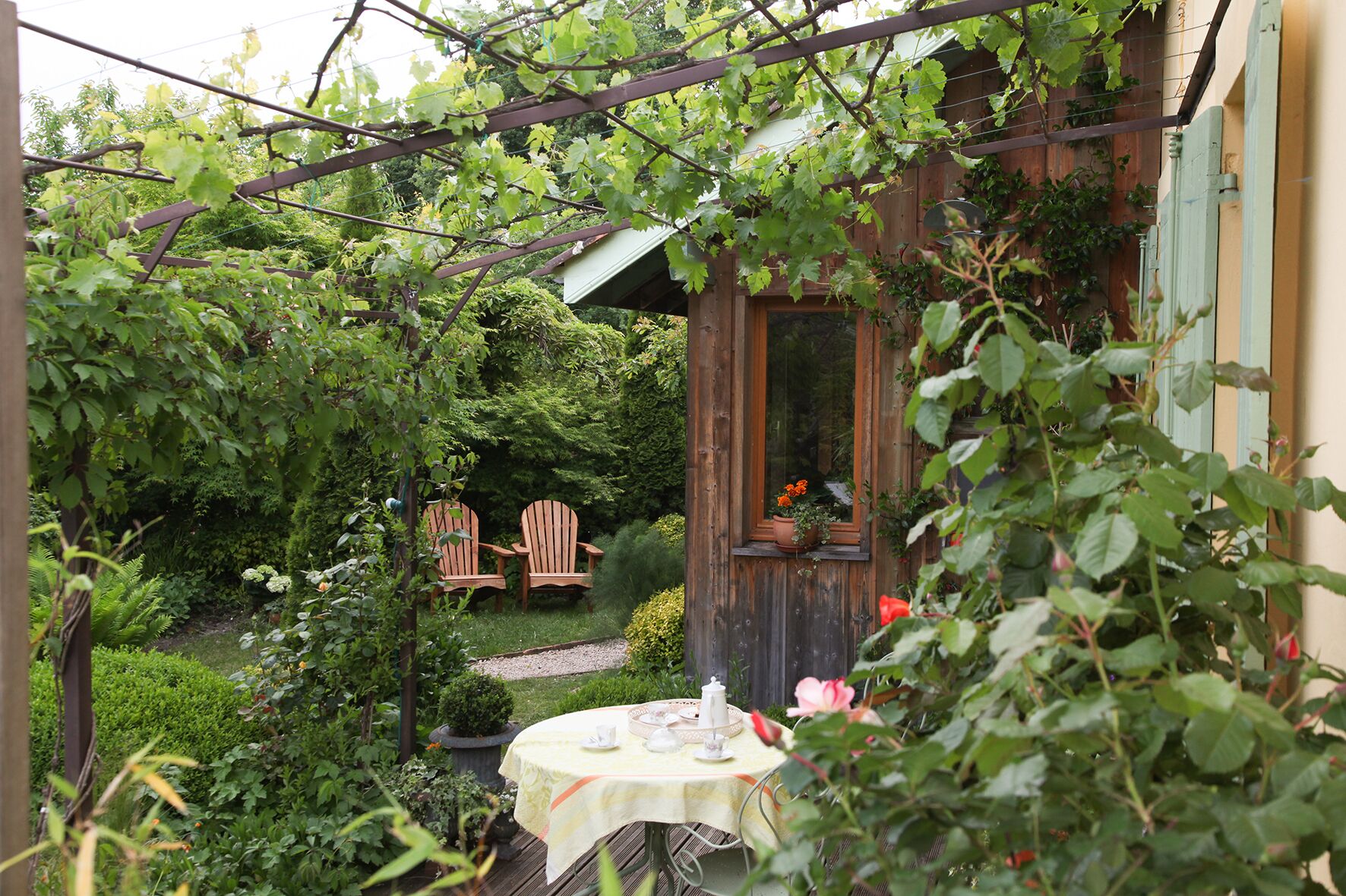 A wooden cottage with a garden, a table with a tablecloth, and two Adirondack chairs.
