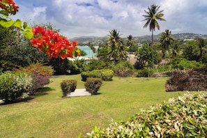 Um jardim bem cuidado com gramados aparados, diversos arbustos e vista clara para uma praia e palmeiras ao fundo.