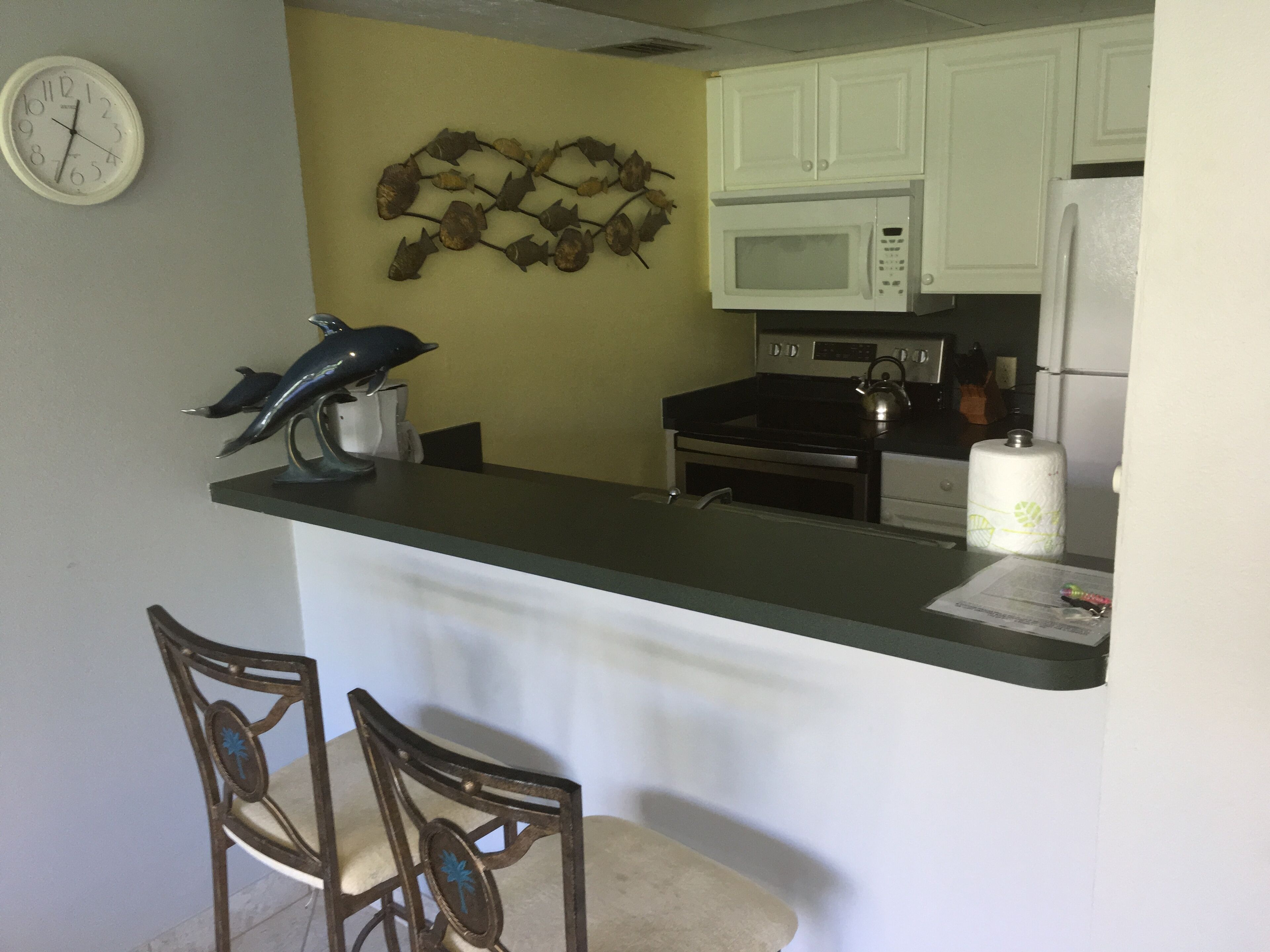 A kitchen with white cabinets, a microwave, a kettle, and a clock on the wall.