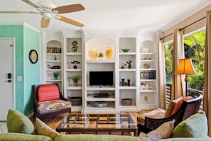 A living room with a TV, bookshelves, a ceiling fan, and a window with curtains.