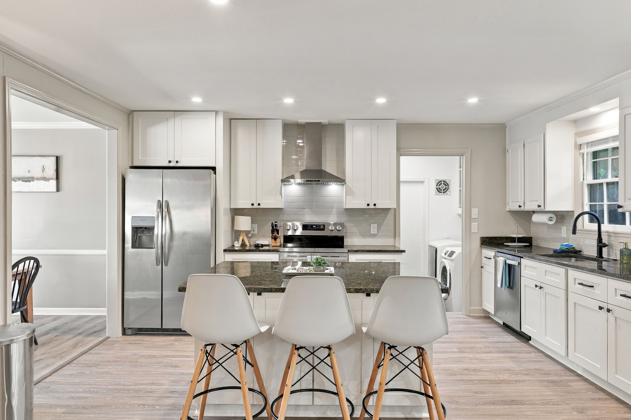 A modern kitchen with white cabinetry, a stainless steel refrigerator, a built-in oven, and a dining area with three white chairs.