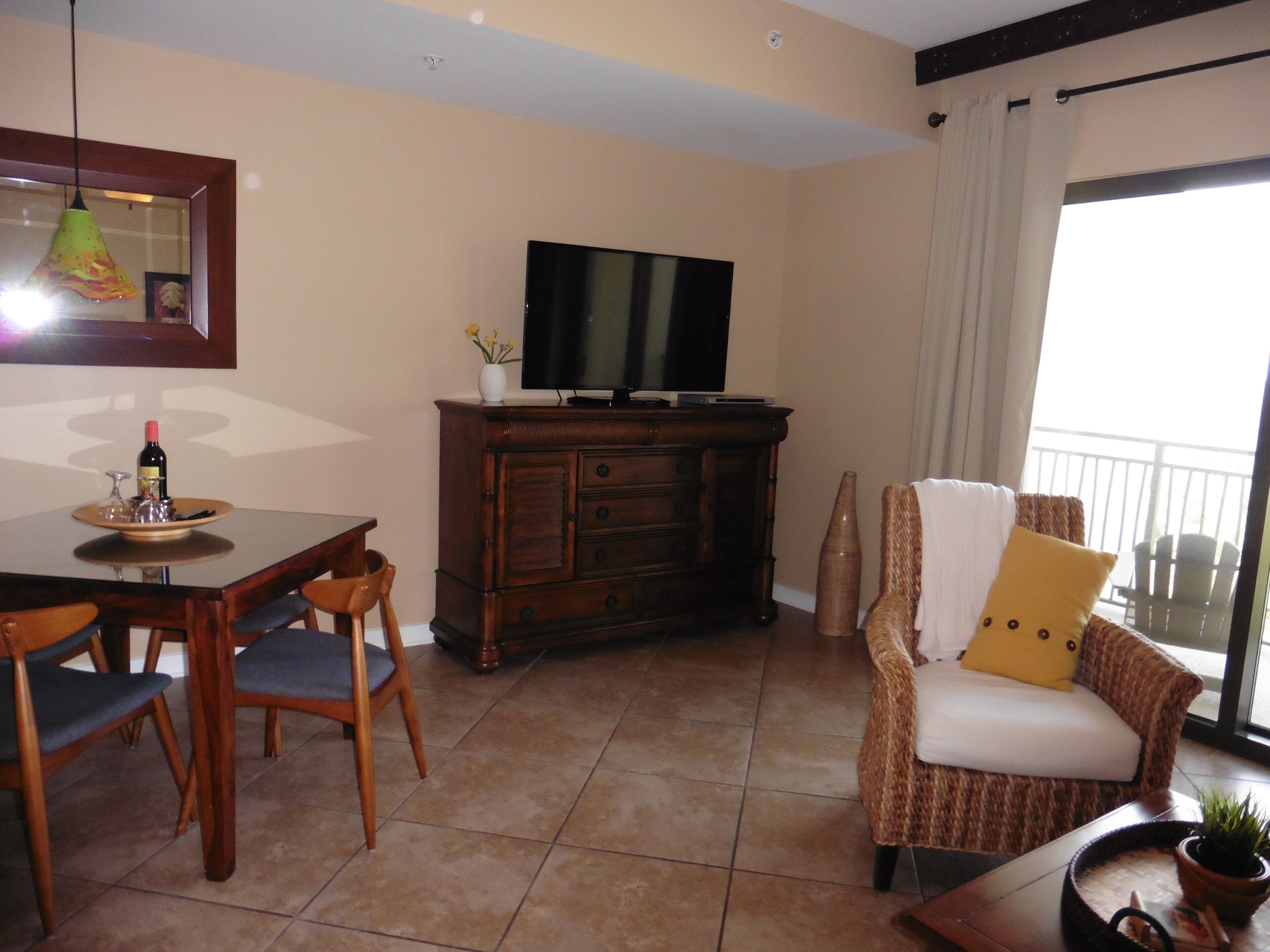 A living room with a TV, wooden furniture, a dining table with chairs, and a balcony with a view.