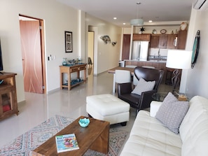 A living room with a sofa, armchair, coffee table, and a magazine on the table. There is a kitchen in the background with a refrigerator and cabinets.