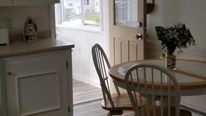 Une cuisine avec des meubles blancs, une table à manger en bois avec des chaises, un ventilateur de plafond et une fenêtre donnant sur des maisons.
