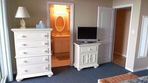 Quarto de hotel com penteadeira branca, televisão em suporte, banheiro com espelho e pia, e porta que dá para outro cômodo.