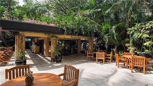 Un área de estar al aire libre con mesas y sillas de madera, rodeada de exuberante follaje y un edificio con un letrero que dice 