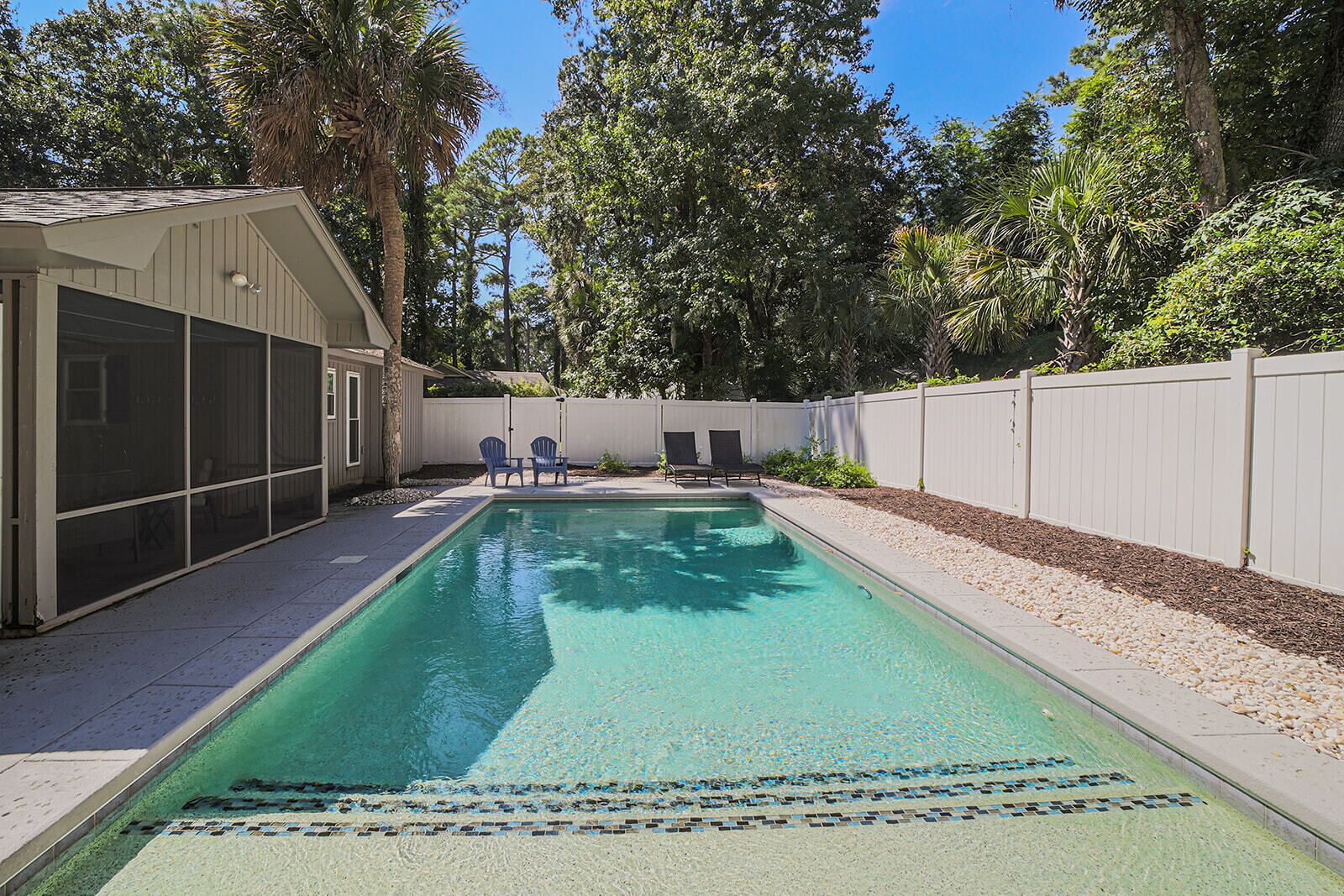 A rectangular swimming pool with clear water, surrounded by a white fence, trees, and a covered patio area with chairs.
