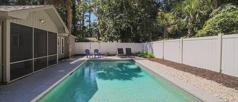 A rectangular swimming pool with clear water, surrounded by a white fence, trees, and a covered patio area with chairs.