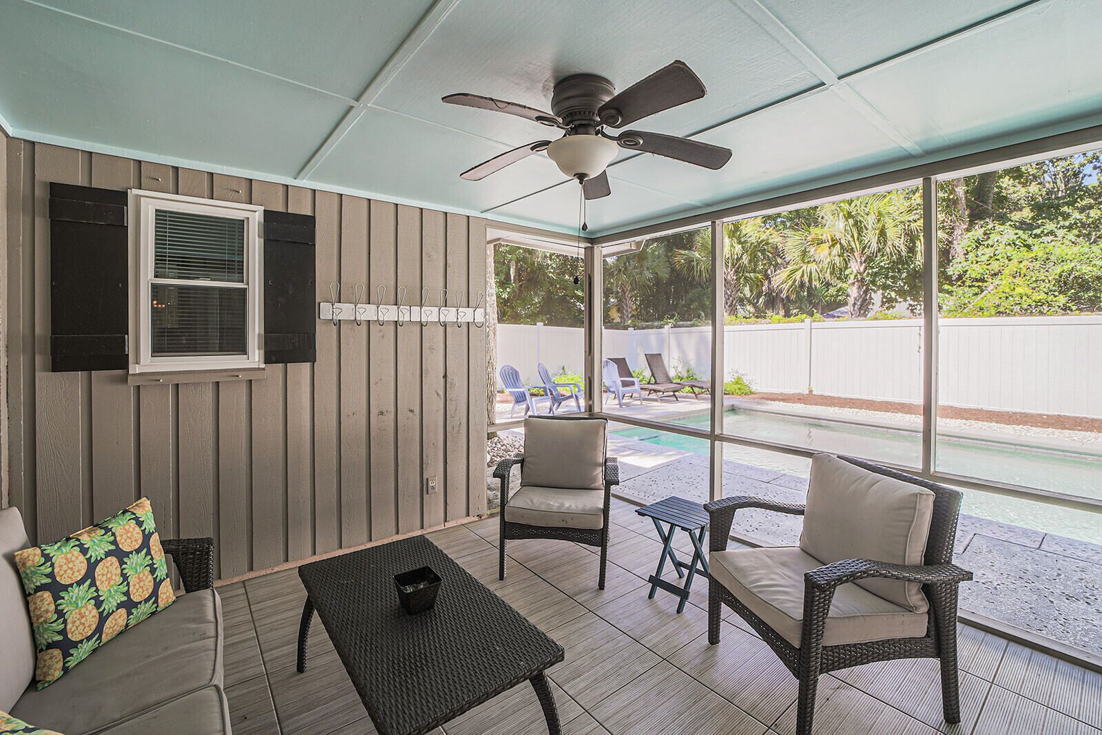 Un patio cubierto con mobiliario de mimbre, un ventilador de techo y vista a un jardín con piscina y sillas.