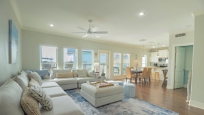 A spacious living room with a sectional sofa, ottoman, and a blue patterned rug. There is a dining area with a table and chairs, and a kitchen in the background.