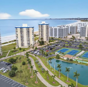 A high-rise hotel with multiple floors, a parking lot, tennis courts, and a body of water.