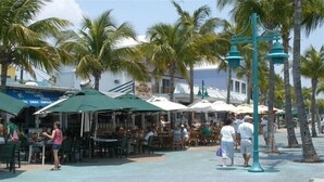 An outdoor dining area with tables and umbrellas, surrounded by palm trees and a building with a sign. - Beautiful condo on the Beach with Heated Pool, Pickleball, free WiFi & parking! (Fort Myers Beach)
