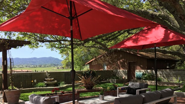 Un'area patio con mobili da esterno bianchi, ombrelloni rossi e vista su un giardino e montagne.
