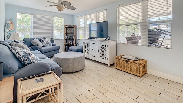 A living room with a grey sofa, a round ottoman, a bookshelf, a television, and a ceiling fan.