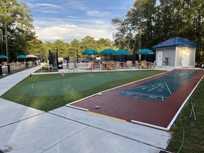 A shuffleboard court with a green and red playing surface, surrounded by outdoor seating and a small building with a blue roof.