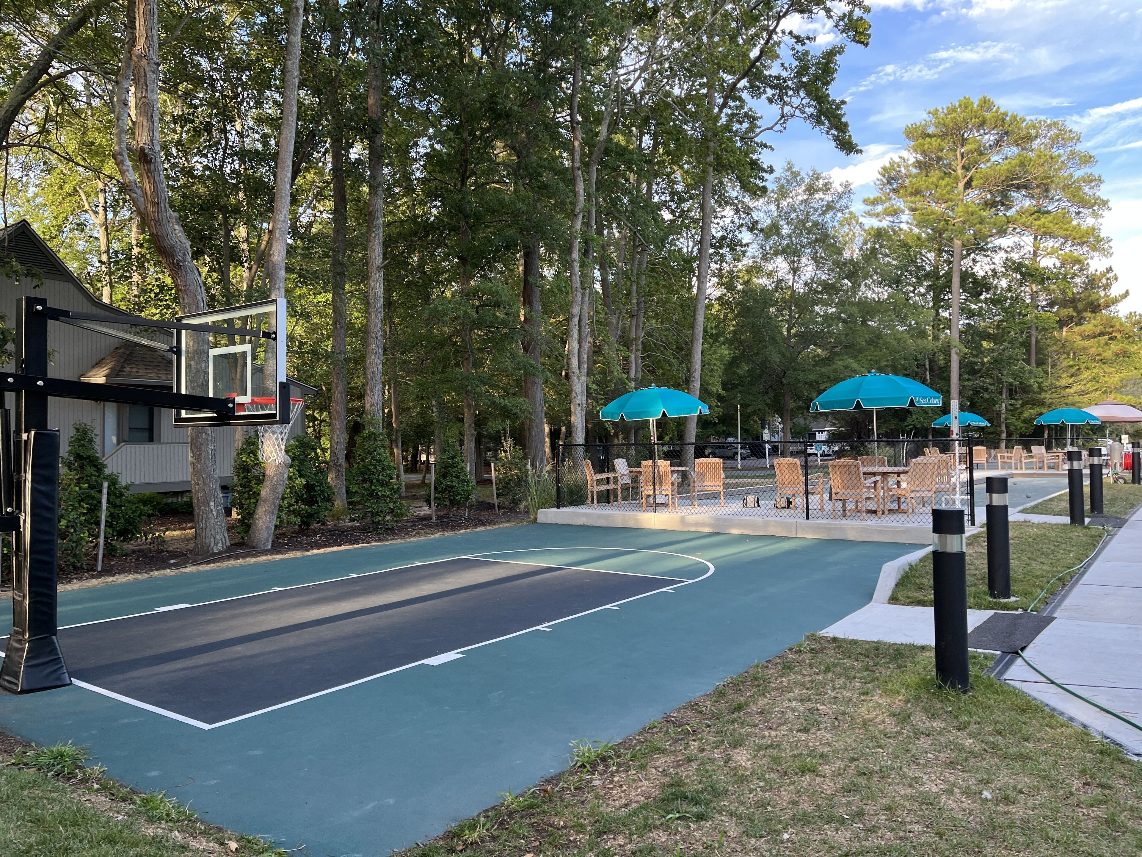 A basketball court with a fence, trees, and outdoor seating with umbrellas.