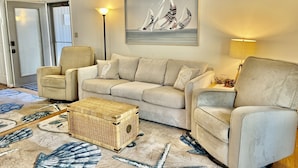 A living room with a beige sofa, two armchairs, a wicker coffee table, and a large painting of sailboats. - Sea Colony West - 2BR, 2BA - 1 Block to Tennis Center, 1/2 Mile to Beach (Bethany Beach)