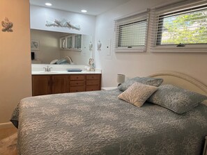 A bedroom with a bed, a nightstand, a window with blinds, and a bathroom visible through an open door. - Sea Colony West - 2BR, 2BA - 1 Block to Tennis Center, 1/2 Mile to Beach (Bethany Beach)