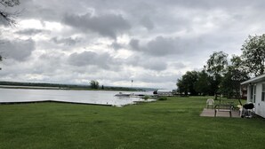 A grassy yard with a dock extending into a lake, a house with outdoor furniture, and a cloudy sky.
