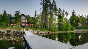 Une jetée en bois s’étendant sur un lac paisible, entourée d’arbres et d’une maison dotée de grandes fenêtres.