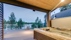 Une terrasse en bois avec un jacuzzi, des chaises et une vue sur un lac entouré d’arbres.