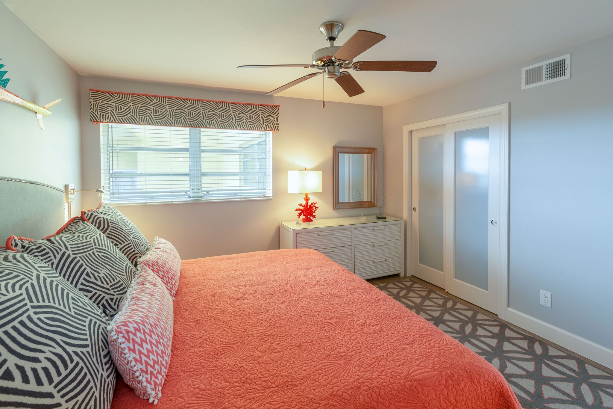 A bedroom with a bed, a ceiling fan, a window with blinds, a bedside table with a lamp, and a dresser.