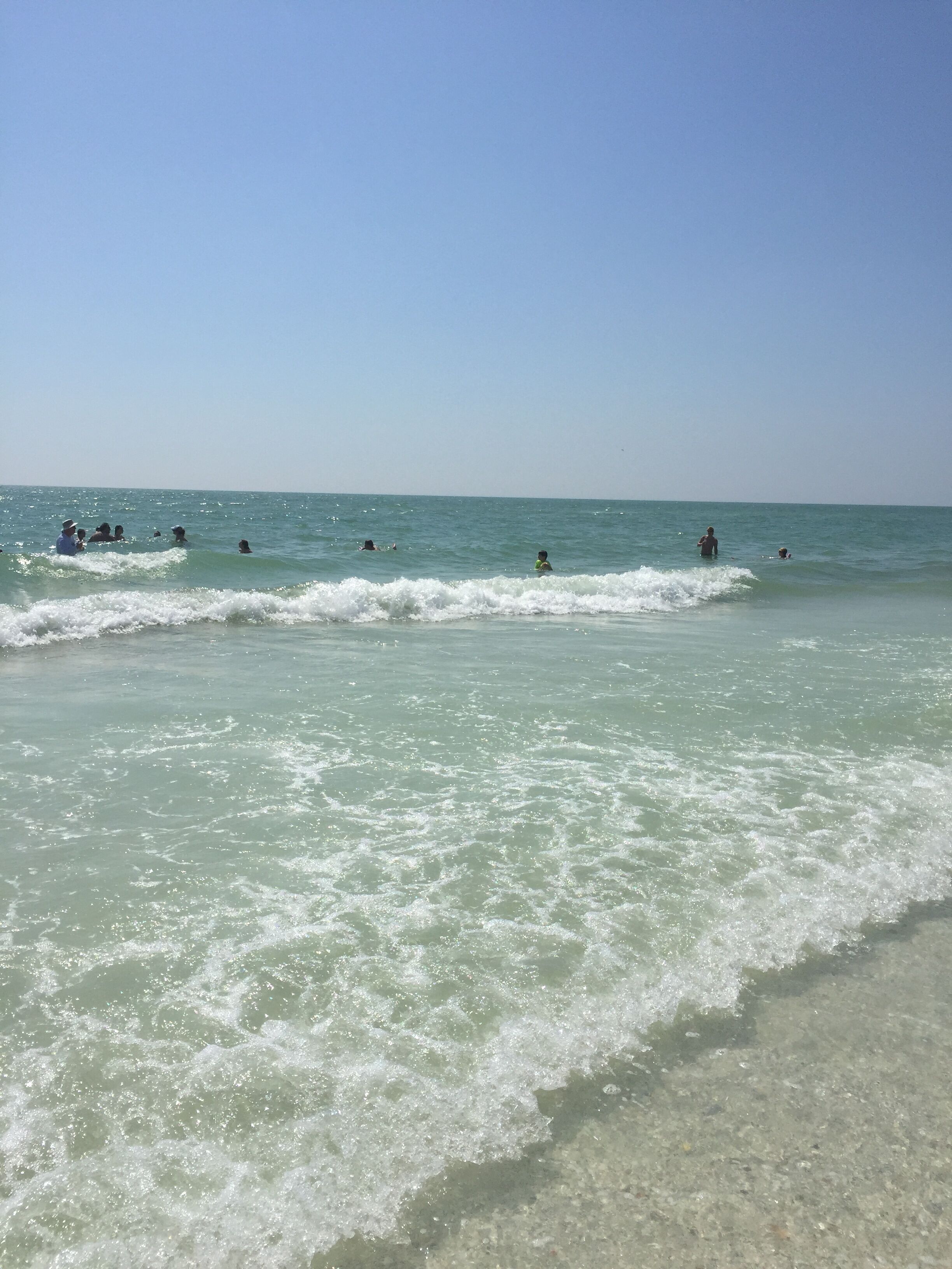 Una playa con agua cristalina y olas, gente nadando y de pie en el agua.