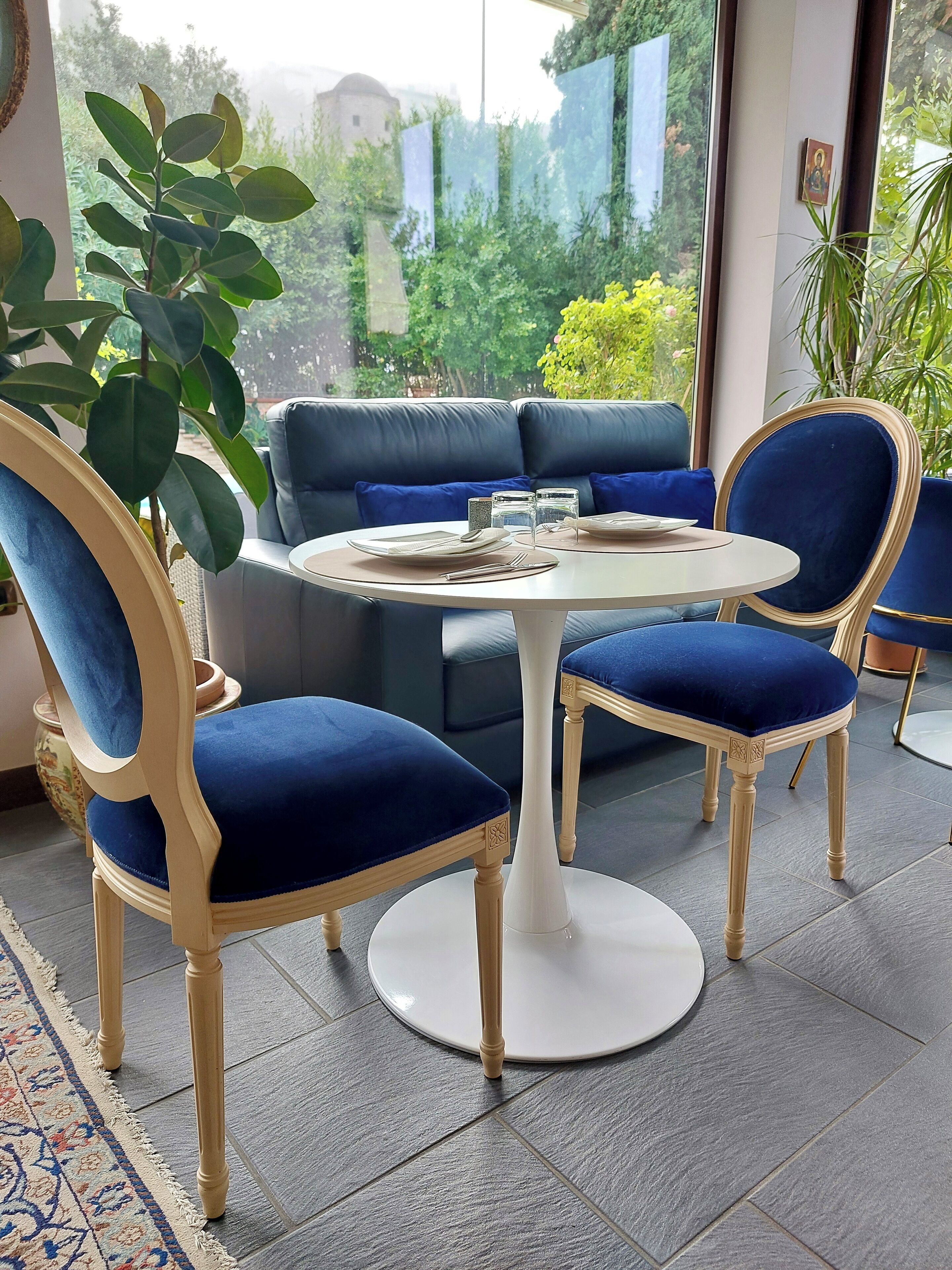 A room with a round table, two blue velvet chairs, a blue sofa, and a view of greenery outside.