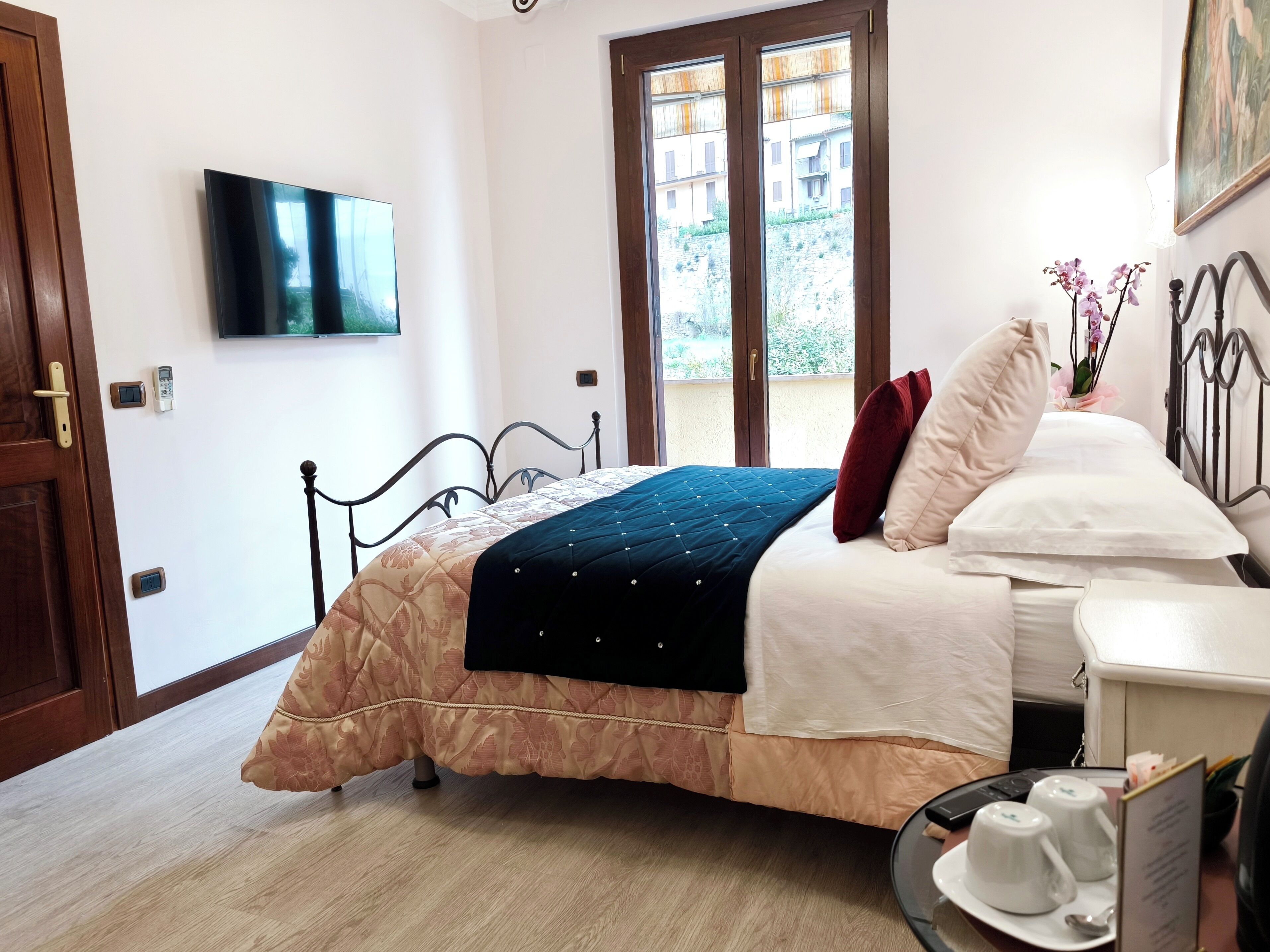 A bedroom with a bed, a TV mounted on the wall, a balcony with a view of buildings, and a small table with cups and a spoon.