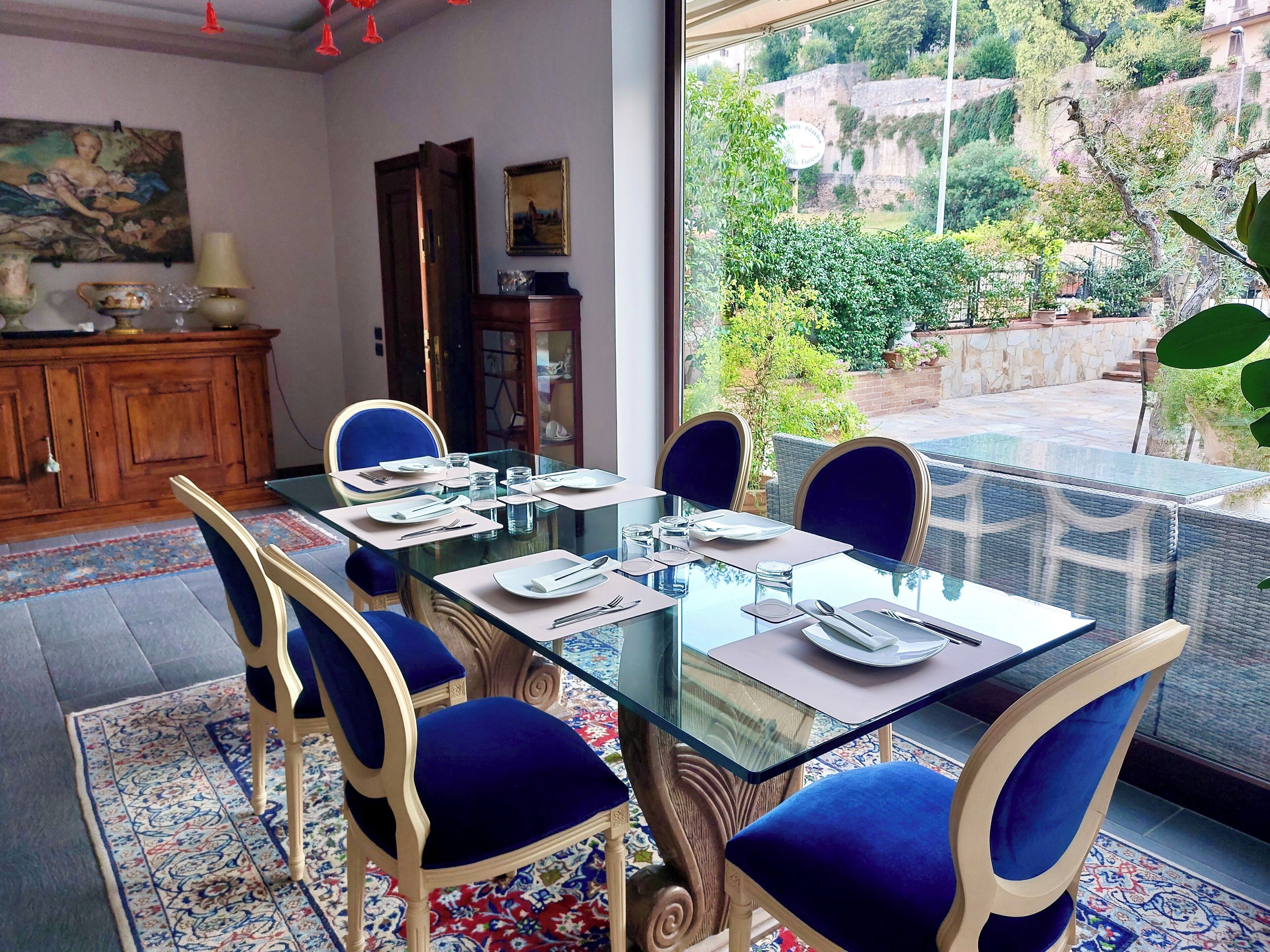Un comedor con una mesa de vidrio, sillas tapizadas en azul y vistas a un jardín y un muro de piedra.