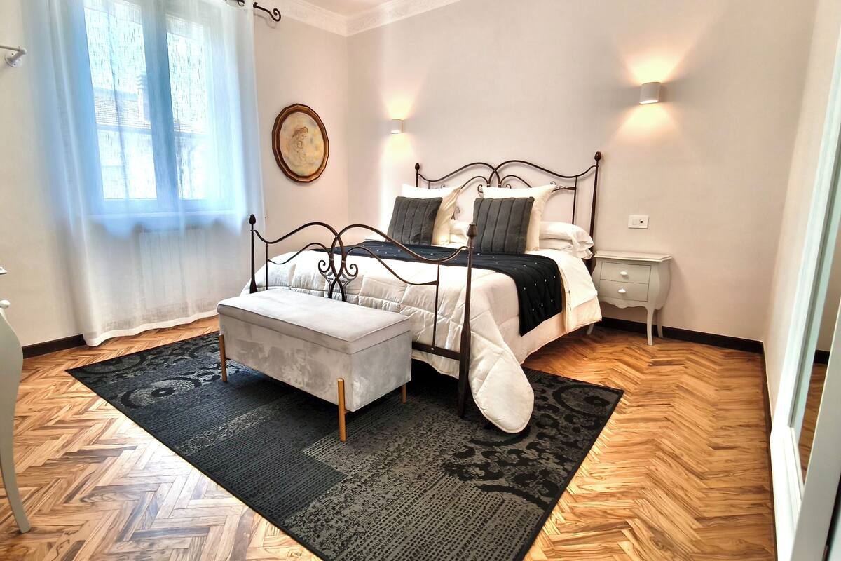 Un dormitorio con cama, mesita de noche y cortinas translúcidas en la ventana.