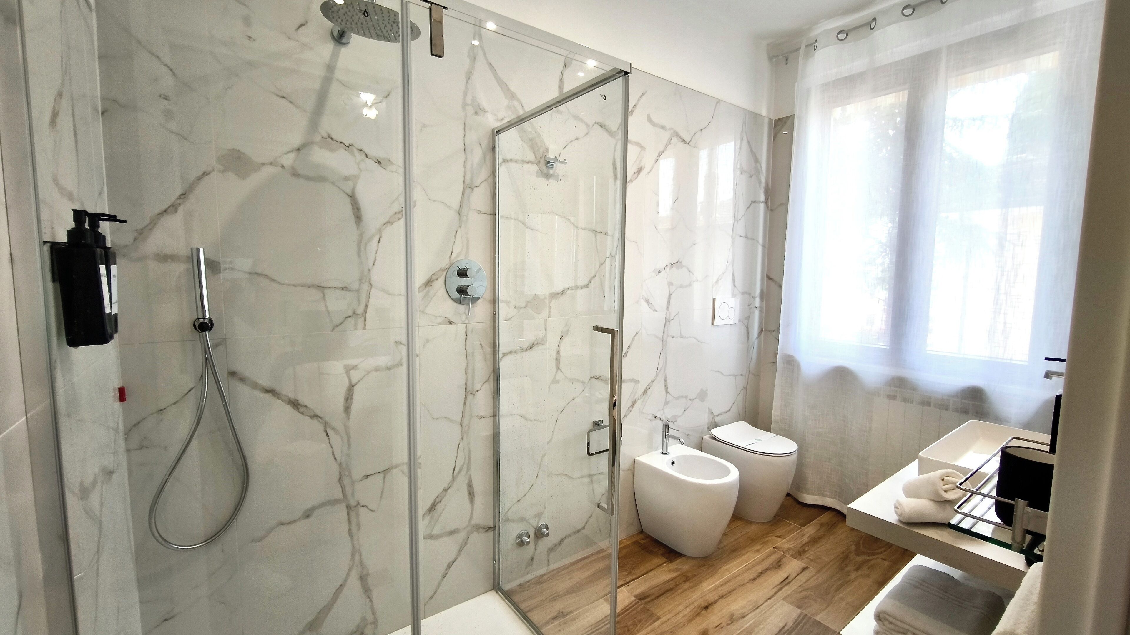 A modern bathroom with a glass-enclosed shower, marble walls, a wooden floor, and a white sink with a towel rack.