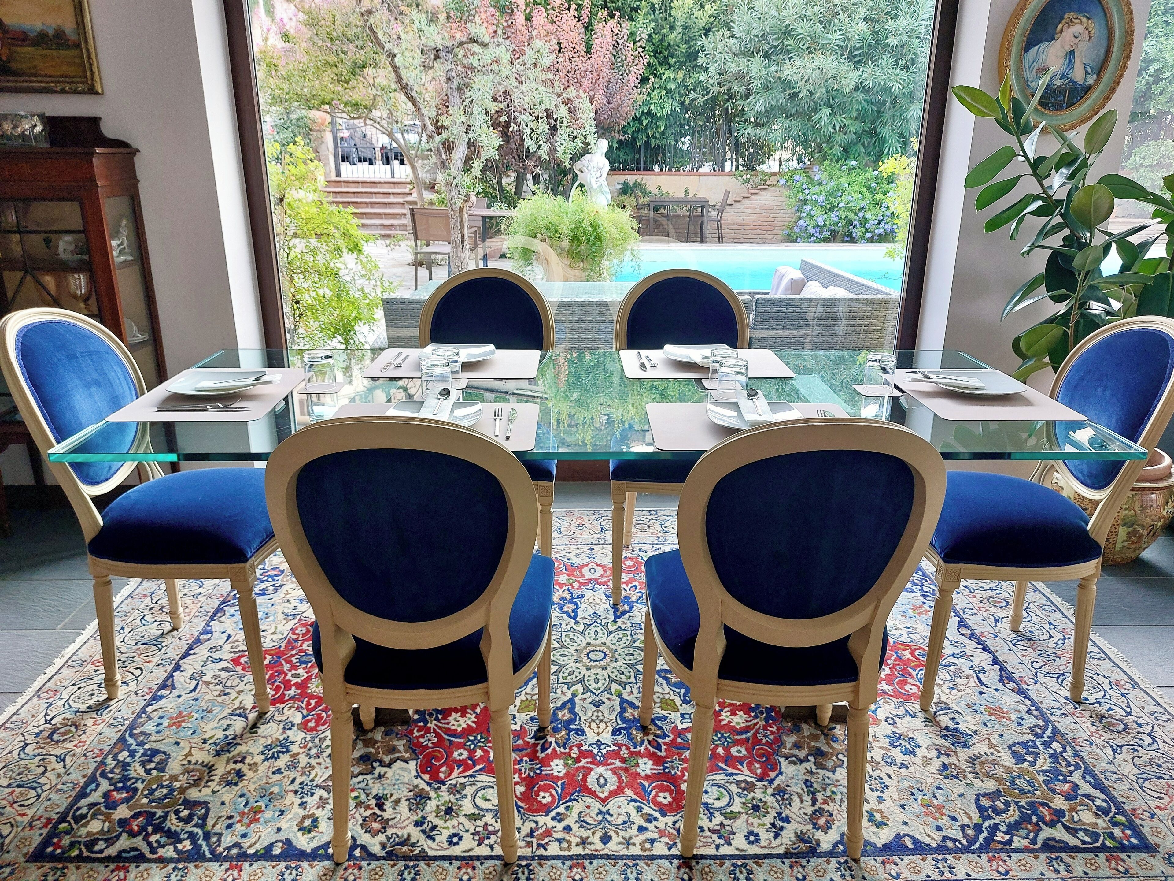 A dining area with a glass table and blue upholstered chairs, a patterned rug, and a view of a garden and pool through large windows.