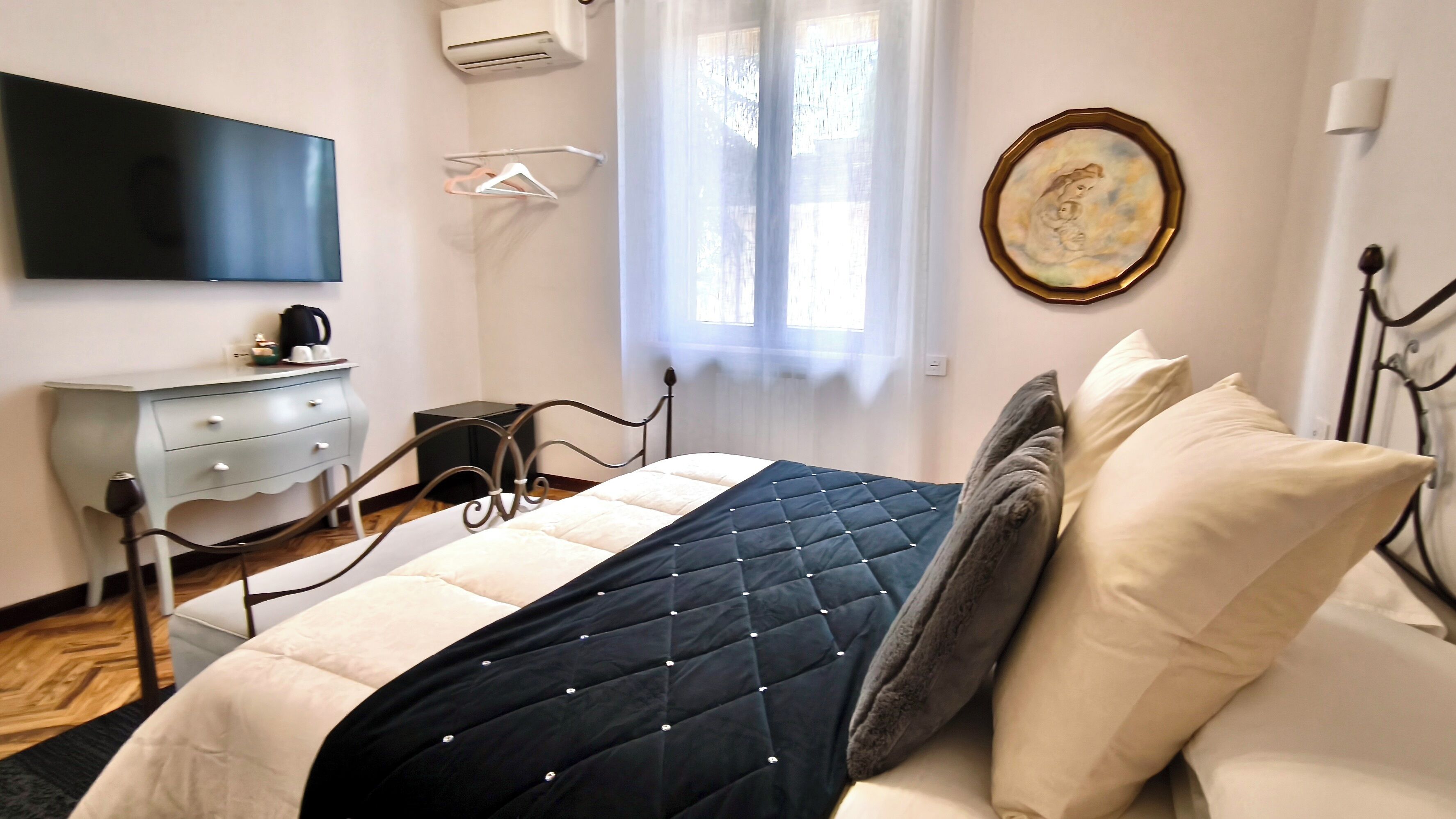 Un dormitorio con cama, mesitas de noche, cómoda, televisor de pantalla plana, ventana con cortinas traslúcidas y un cuadro colgado en la pared.