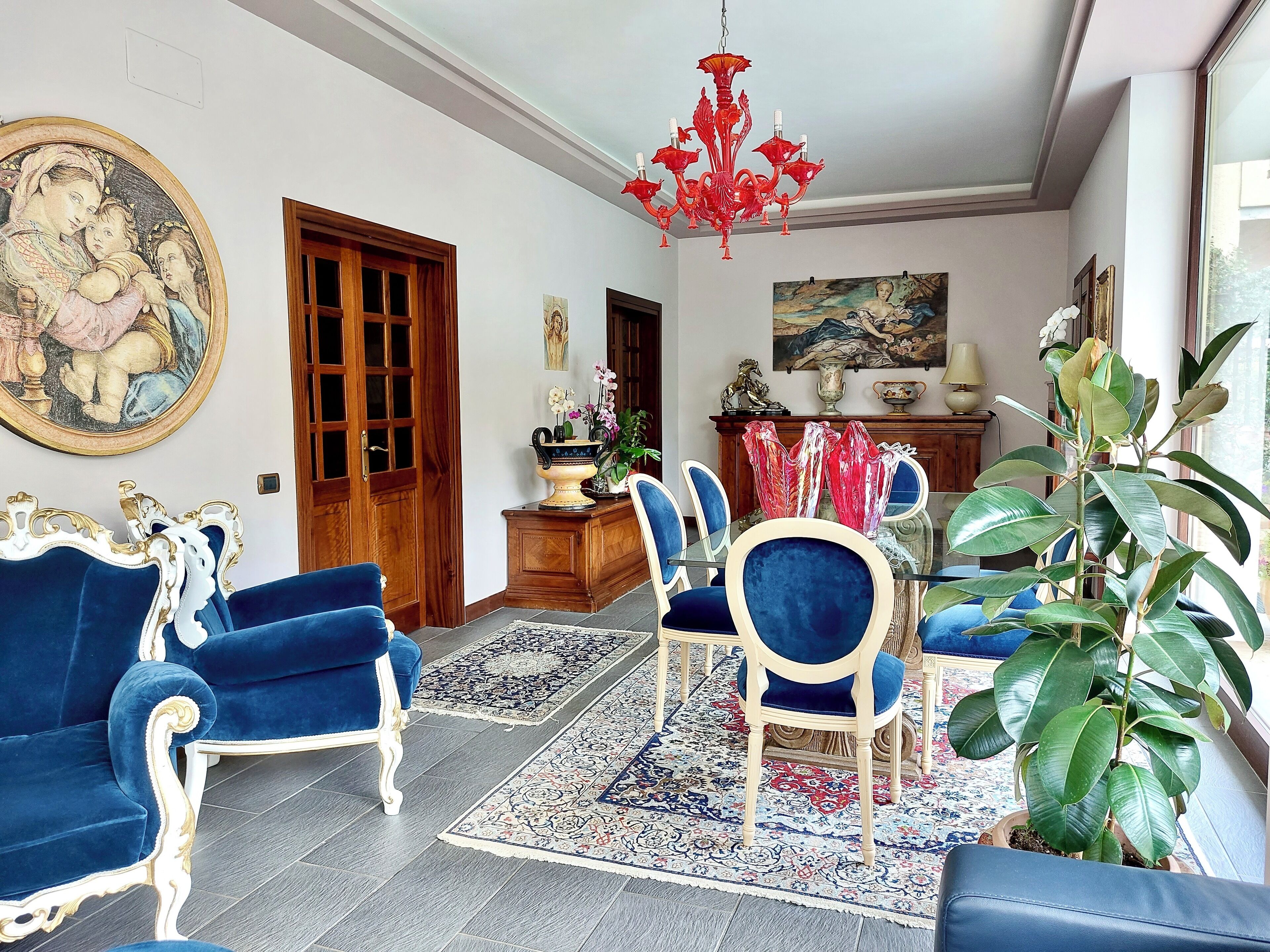 A room with a dining area, a large painting, a chandelier, and a potted plant.