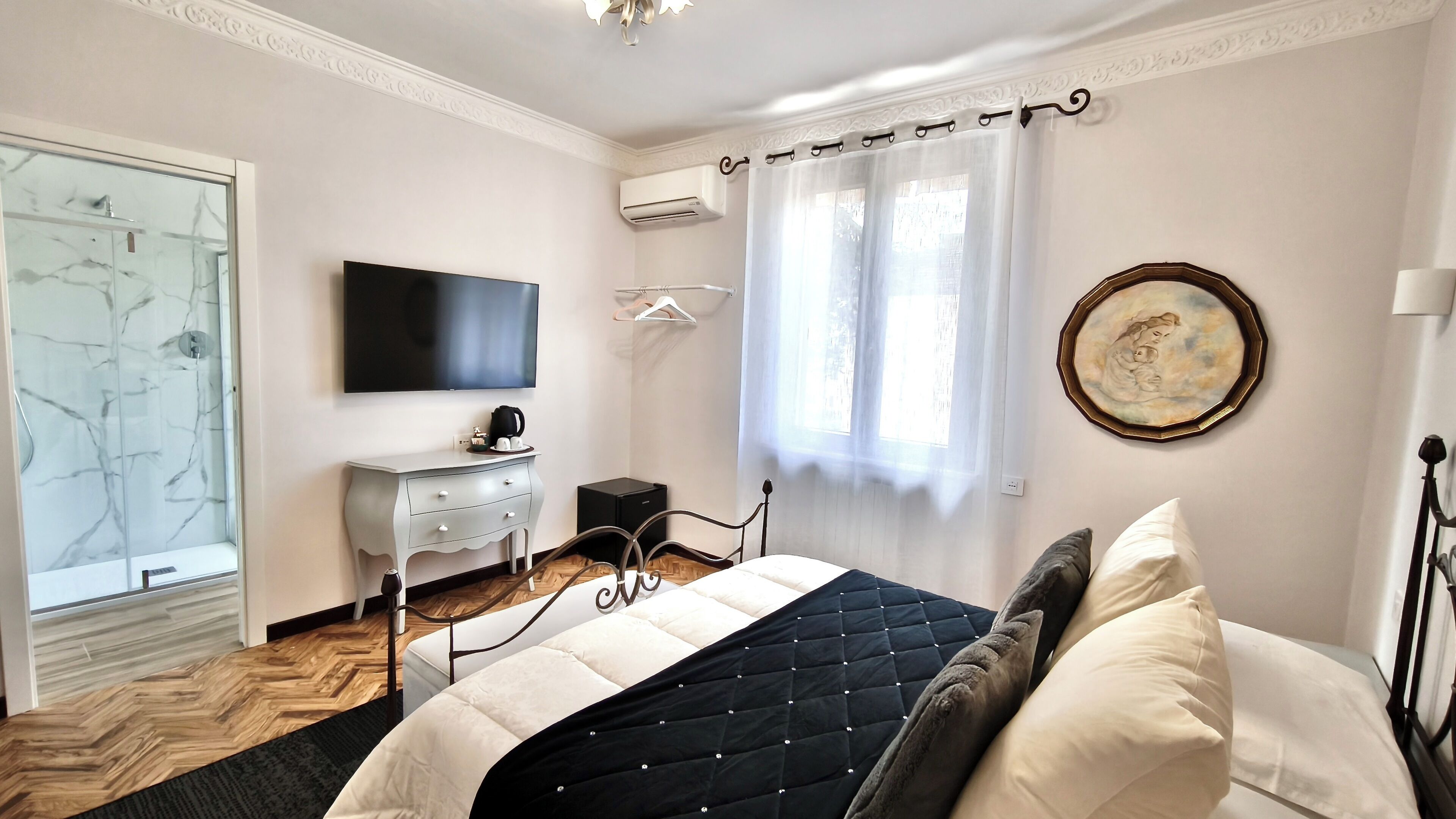 A bedroom with a bed, a TV, a dresser, a shower, and a window with curtains.