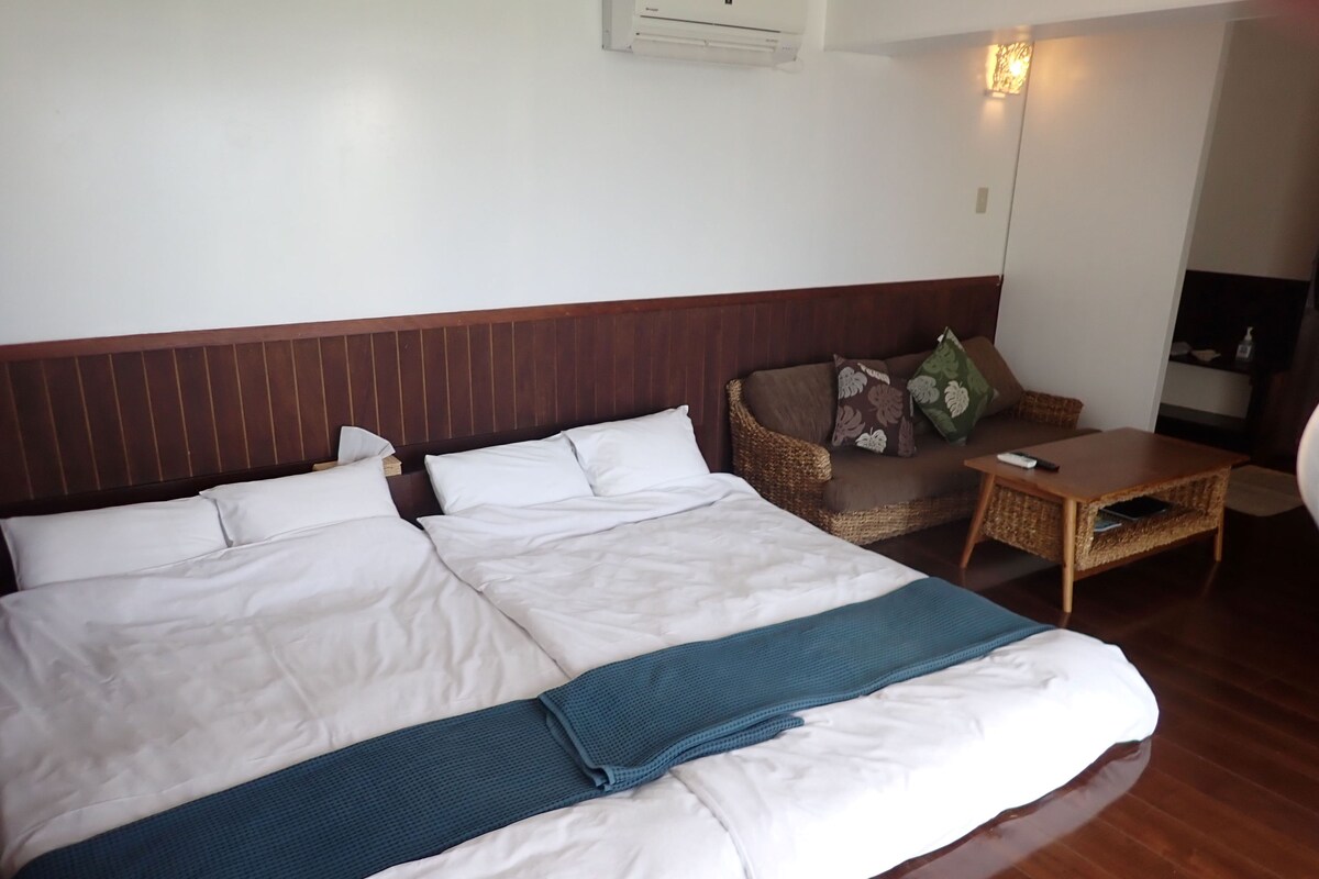 A hotel room with a large bed, a wooden headboard, a sofa, a small table, and a wall-mounted air conditioner.